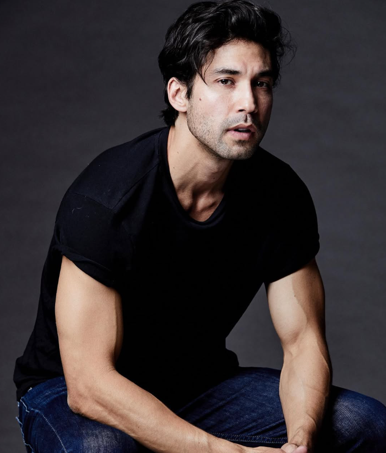 An attractive mid-30's male in black shirt and blue jeans sits leaning forward before a dark gray background.