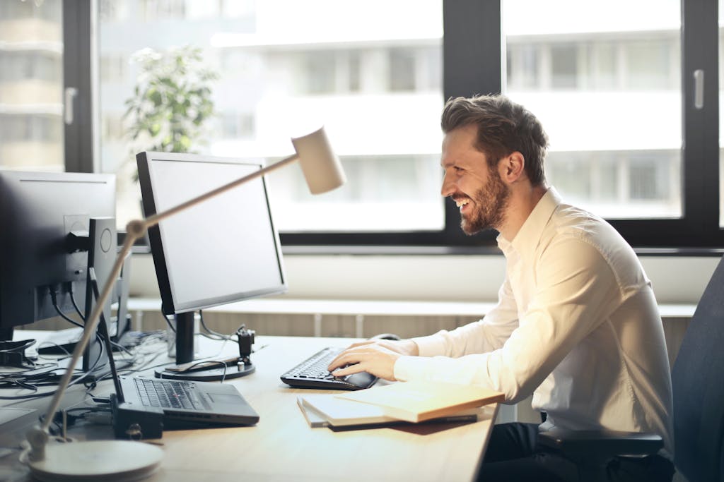 A man smiling, working at an office desk on marketing strategy with a computer and natural daylight streaming in through large windows.