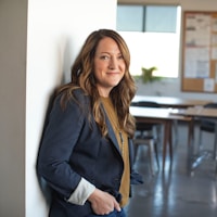 A woman in a modern suit-jacket smiles for a portrait in a professional school environment setting.