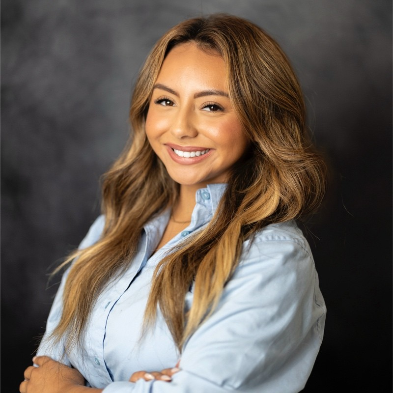 Professional headshot of a smiling woman with long light-brown hair, wearing a light blue button-down shirt, standing with arms crossed against a dark studio background