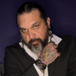 Portrait of a man with slicked-back dark hair and a gray beard, wearing a black suit and white shirt, with facial piercings, large earrings, and a tattooed hand resting near his collar against a dark background.